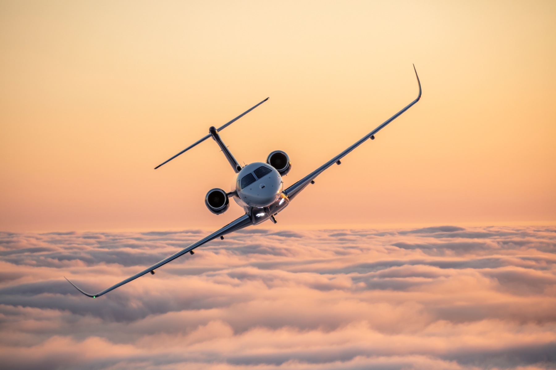A white business jet with dark engines flies head-on over clouds during sunset.